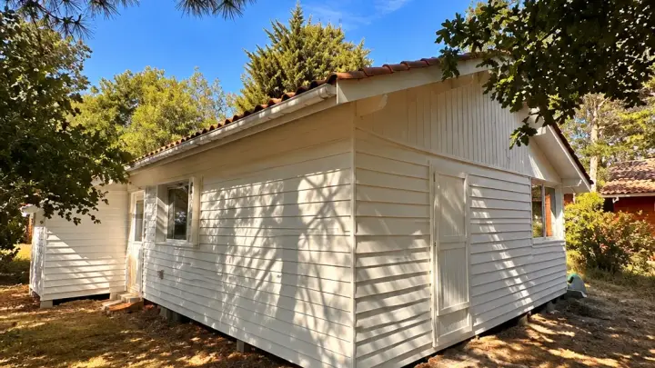 Arrière de la maison, bardage horizontal repeint en blanc cassé, à l’ombre des pins