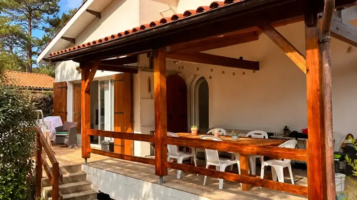 Terrasse couverte aux poteaux, poutres et garde-corps en bois de teinte chaude, devant une maison blanche